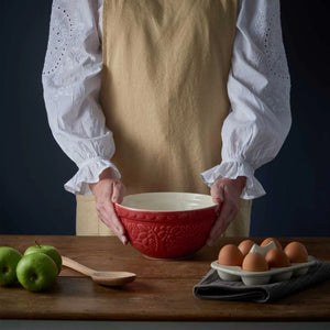 Person holding the Mason Cash In The Forest Hedgehog Red Mixing Bowl on a wooden table with apples, eggs, and a wooden spoon.