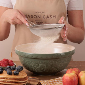 Mason Cash In The Forest Owl Mixing Bowl in Dark Green (26 cm) styled next to fruit and pancakes, with a person sifting flour into the bowl.