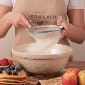 Person sifting flour into a Mason Cash In The Forest Owl Mixing Bowl with fruits and pancakes on a table.