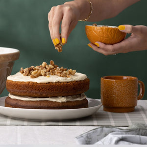 Person garnishing a cake with walnuts next to a mug and holding the mason cash in the forest prep bowl.