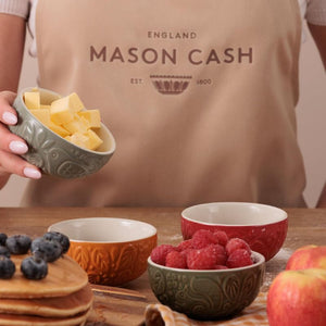 Person holding a bowl of butter with Mason Cash apron, surrounded by the in the forest prep bowls and fruits on a wooden surface.