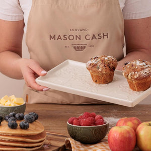 Person holding the Mason Cash In The Forest Serving Platter with muffins, surrounded by fruits and pancakes, wearing a Mason Cash apron.