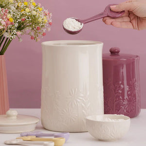 Person measuring flour with a spoon over the Mason Cash In The Meadow Storage Jar in Cream on a pink background