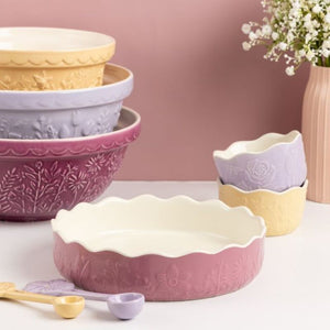 A set of pastel-colored ceramic bowls and measuring cups with embossed floral patterns by Mason Cash, arranged on a white surface against a dusty pink background.