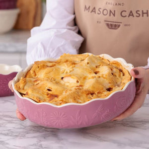 A person holding the Mason Cash In The Meadow Pink Pie Dish with a scalloped rim and embossed floral pattern, filled with a baked apple pie on a marble countertop.