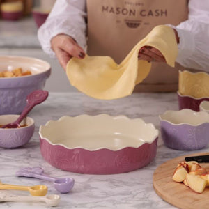 A person wearing a tan apron holds a pastry sheet over a scalloped the Mason Cash In The Meadow Pink Pie Dish. The scene shows a coordinated set of pastel purple and cream ceramic baking dishes and measuring spoons on a marble countertop, with sliced fruit in a cutting board.