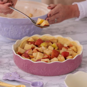 Mason Cash In The Meadow Pink Pie Dish filled with unbaked apple pie crust and diced apples, with a person's hands using a spoon to transfer fruit from a mixing bowl in the background.