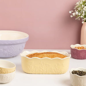 Mason Cash In The Meadow Yellow Loaf Pan next to coordinating ceramic bowls on a pink background.