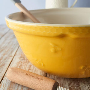Close up view of the Mason Cash Sweet Bee Yellow Mixing Bowl with whisk and wooden spoon on rustic wooden surface - embossed bumblebees and honeycomb detail shown.