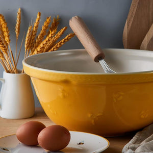 Mason Cash Sweet Bee Yellow Mixing Bowl with whisk, eggs, wheat stalks and breadboard on kitchen table.