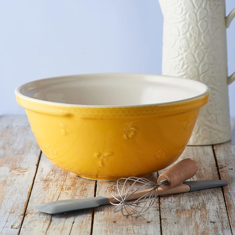 Mason Cash Sweet Bee Yellow Mixing Bowl with embossed bumblebee and honeycomb detail, shown with whisk and spatula in front on a rustic wooden table.