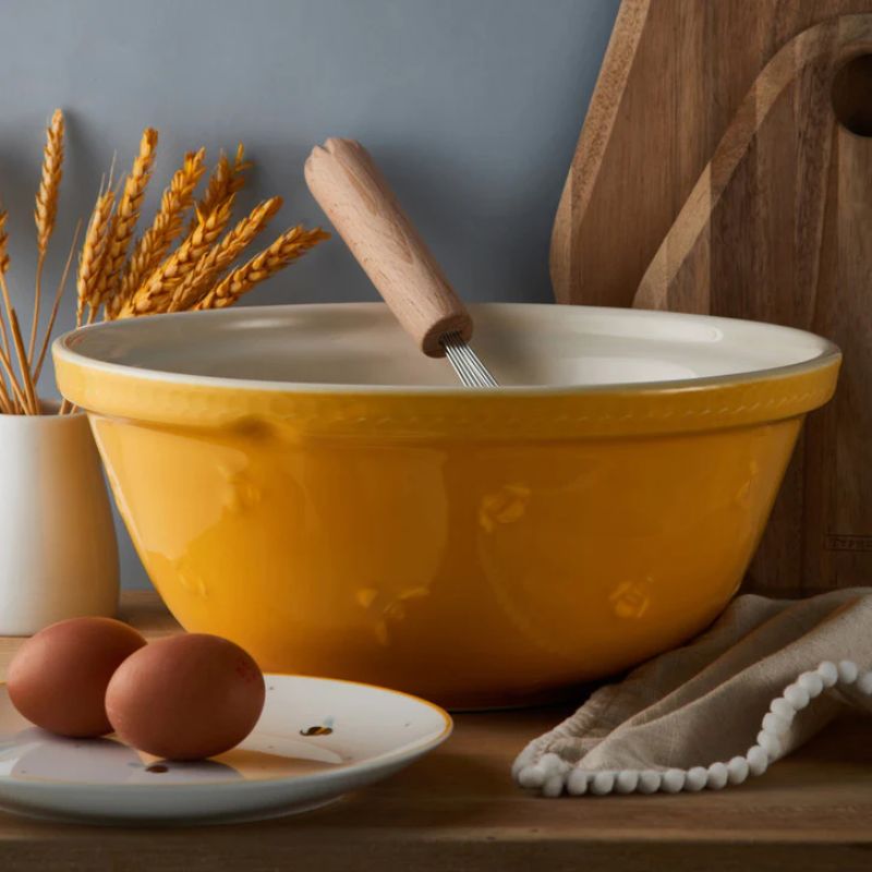Mason Cash Sweet Bee Yellow Mixing Bowl styled with eggs, whisk and wheat stalks on rustic table.