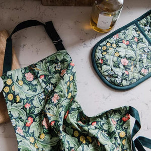 Green floral patterned apron and oven mitt from the Museums & Galleries Leicester Wallpaper range shown on a kitchen bench next to a bottle of olive oil.
