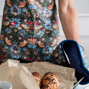 Person wearing a Museums & Galleries Strawberry Thief apron with a tray of pastries and a cup on a table.