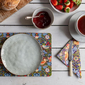 Breakfast setting with a plate, knife, and William Morris Strawberry Thief placemat and paper napkin on a wooden table.
