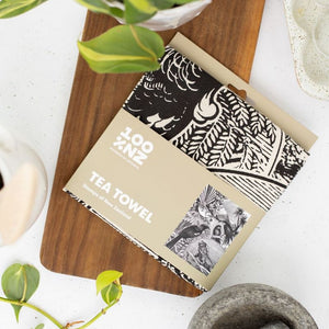 Black and white NZ Stamp tea towel by 100%NZ shown in retail packaging in a kitchen scene.