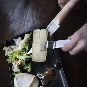 Person slicing cheese with Opinel cheese knife and fork.