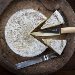 Opinel cheese knife and fork slicing into a round cheese on wooden board.