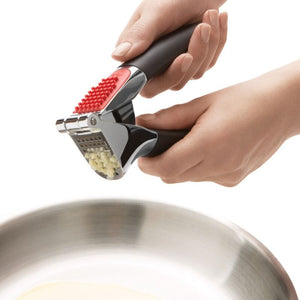 Hand using the OXO Good Grips Garlic Press to crush garlic over a bowl on a white background
