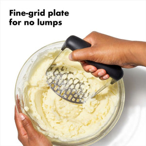 Hand using the OXO Good Grips Smooth Potato Masher on mashed potatoes with text 'Fine-grid plate for no lumps'.