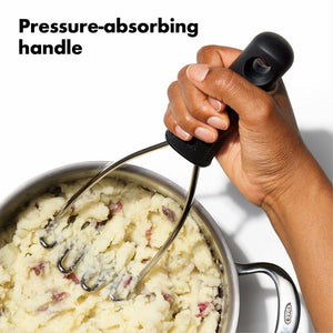 A hand holding a the OXO Good Grips Wire Potato Masher over a pot of mashed potatoes, with the text 'Pressure-absorbing handle' on a white background.