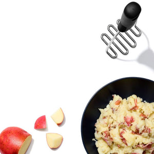 Mashed potatoes in a black bowl with a potato masher and sliced red potatoes on a white background