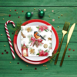 A festive holiday place setting on a green wooden table, featuring a the PAW Paper Napkins in the Bullfinch with Lampion design layered on a red and white plate. A red-and-white striped candy cane, gold flatware, and Christmas ornaments surround it.