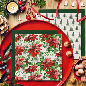 PAW Paper Napkins in the Glory Poinsettia pattern layered on a dark green napkin on a red plate, surrounded by acorns, ribbon, and Christmas berries.