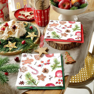 A styled holiday tabletop scene showing the PAW Paper Napkins in the Homemade Sweets pattern. The napkins are arranged on wood slices with red napkins underneath, alongside meringue, star-shaped cookies, a mug, and fruit in a bowl.