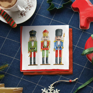 PAW Paper Napkins in the Nutcrackers Soldiers design, placed on a red patterned napkin over a blue plaid tablecloth with Christmas-themed props.