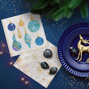 A styled flat-lay of PAW Paper Napkins Peacock Collection design on a dark blue background with pine branches, gold confetti, string lights, and a navy plate with a gold reindeer decoration.