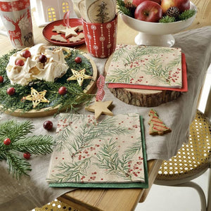 A styled holiday tabletop scene featuring the PAW Paper Napkins in the We Care Sprigs Holly design, layered with red and green napkins. The setting includes a white cake with whipped cream and cranberry garnish on a wooden platter, a red mug with a snowflake pattern, a bowl of apples, and wooden star decorations.