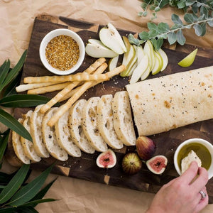 Peer Sorensen End Grain Cutting Board with sliced bread, figs, pears, and a wrap, surrounded by greenery.