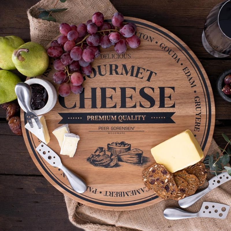 Peer Sorensen round wooden gourmet cheese board with cheese, grapes, and utensils on a rustic wooden surface.