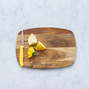 Peer Sorensen Slim Line Rounded Cutting Board on a marble table with a knife and chopped vegetables.