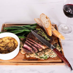Carving board topped with sliced steak, bread, vegetables, and a bowl of sauce.