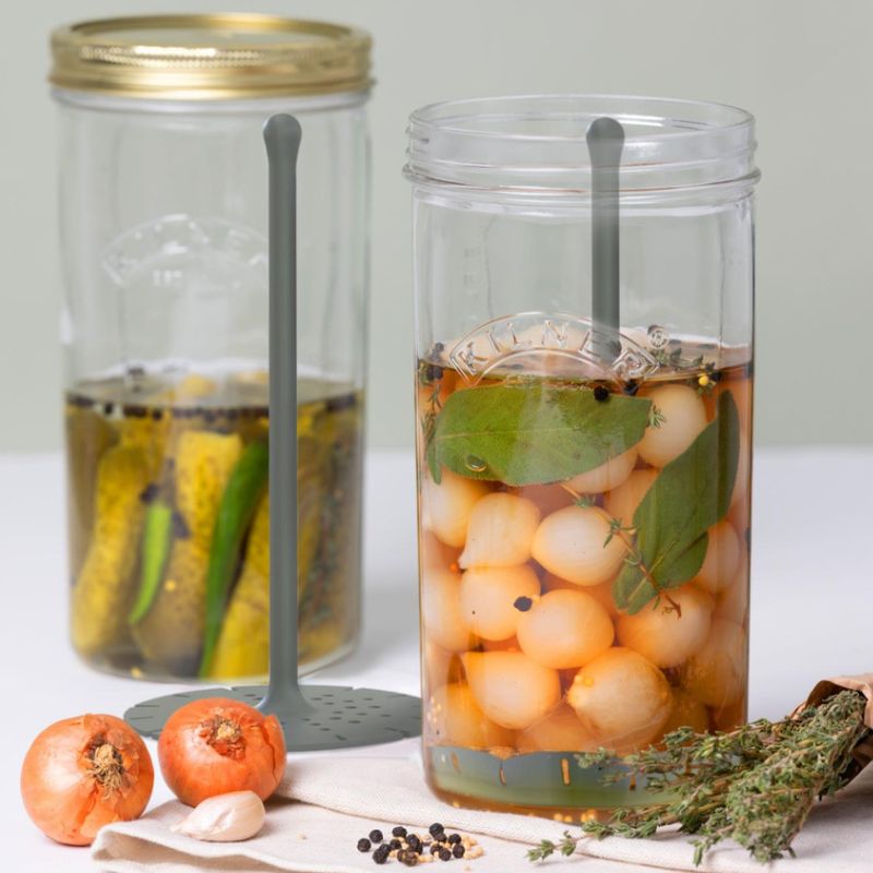Two Kilner Pickle Jars with Lifters filled with gherkins and pickled onions, styled with herbs and vegetables.