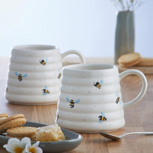 Two Price & Kensington Sweet Bee Hug Mugs with a ribbed beehive design and printed bumblebee motifs, displayed on a wooden table with cookies, honey, and a spoon in a styled setting.