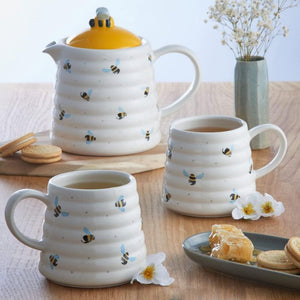 Price & Kensington Sweet Bee Hug Mug with a ribbed beehive design, next to a white teapot with a yellow lid, and styled with cookies, honey, and dried flowers on a wooden table.