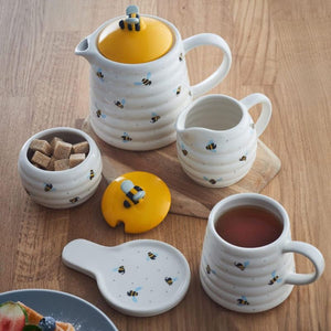 A ceramic tea set by Price and Kensignton arranged on a wooden table, including a white ribbed teapot with a yellow lid, a matching jug, a small bowl of sugar cubes, a cup of tea, and a spoon rest, all decorated with small bumblebee motifs.
