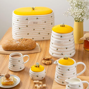 Price & Kensington Sweet Bee collection featuring white ribbed containers with sweet bee designs, bright yellow lids and matching small cups and bowls, styled on a wooden table with bread, cookies, honey, and flowers.