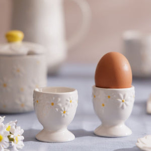 Price & Kensington Daisy Egg Cup Set with embossed daisy flowers and yellow centers, one holding a brown egg and the other empty, set on a light blue tablecloth with a softly blurred breakfast scene in the background.