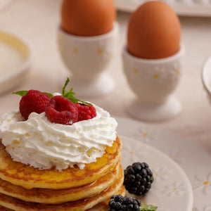 A close-up of a stack of golden pancakes topped with whipped cream, fresh raspberries, and mint, served on a plate with blackberries, while two brown eggs sit in the Price & Kensington Daisy Egg Cups.