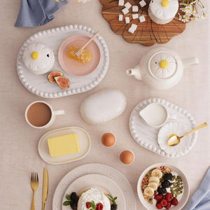 A styled breakfast table setting with multiple white ceramic dishes, plates, a teapot, cups, butter, fruit, eggs, and a small teardrop-shaped container from the Price & Kensington Daisy collection, arranged on a light beige surface.