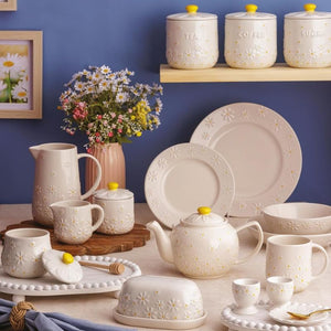 A coordinated ceramic tableware set from the Price & Kensington Daisy collection, arranged on a tabletop against a blue wall, including plates, cups, a teapot, pitcher, bowls, and canisters with daisy embossing and yellow lids.