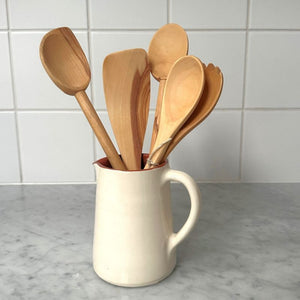 Roger Orfèvre olive wood kitchen utensil collection shown in a terracotta jug on a kitchen bench.