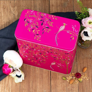 A bright pink rectangular metal tin with decorative botanical and bird illustrations, styled on a wooden surface with flowers and nuts around it.