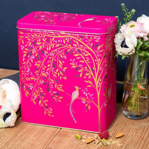 Sara Miller Chelsea Larder Tin, decorated with gold and green leafy branches, small birds, and white flowers. The tin sits on a wooden tabletop next to a glass vase with pink flowers, with a dark blue wall in the background.