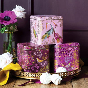Three Sara Miller Haveli Garden Square Tin Caddies with bird illustrations on a wooden surface with flowers and a purple background.