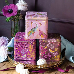 Three Sara Miller Haveli Garden Square Tin Caddies with floral and bird illustrations are stacked on a gold tray. The top box is light pink with pink flamingos, while the two boxes below are deep purple and burgundy with purple and gold floral patterns.